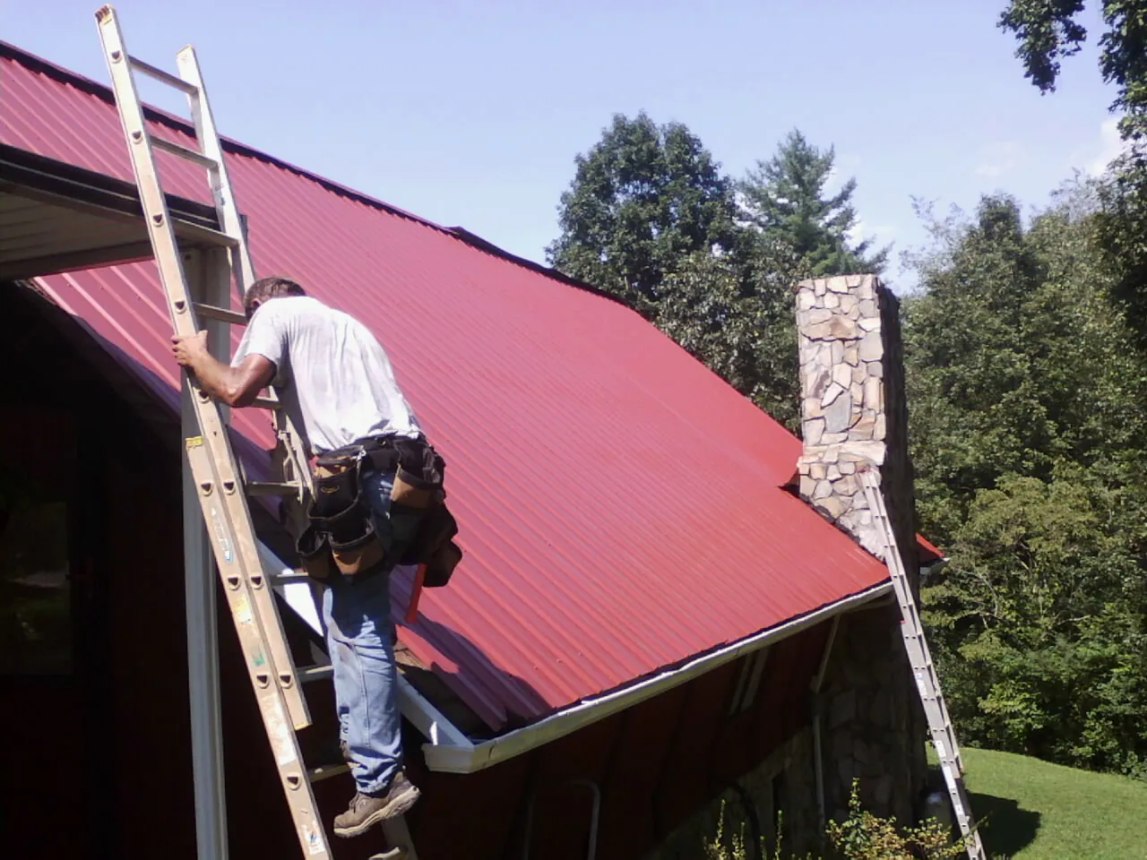 Roofer on a ladder beside a red metal roof and stone chimney — Soffit & Fascia Repair by Roofing Brothers in Munford